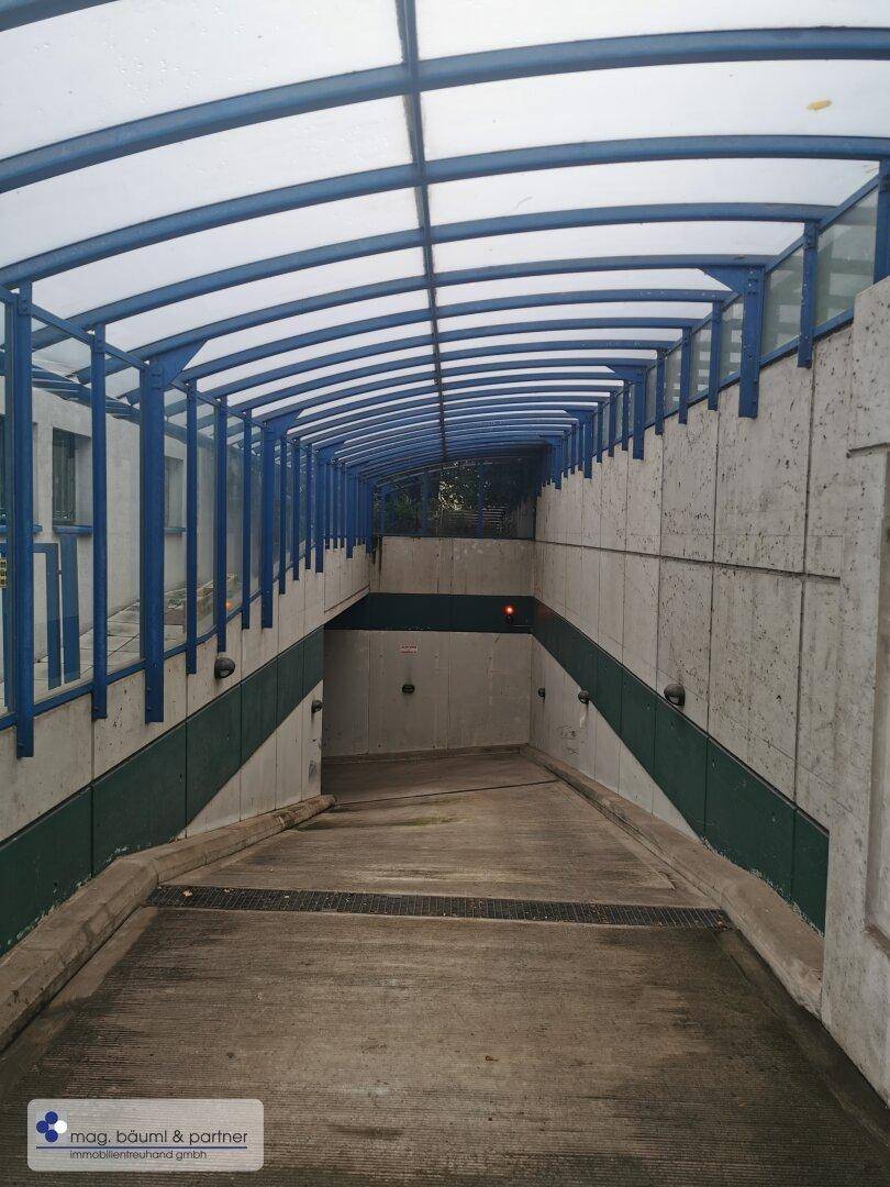Überdachte Rampe führt hinab in die Tiefgarage, mit blauen Geländern und Betonwänden.