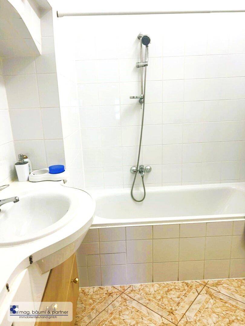 Badezimmer mit Badewanne, Duscharmatur und Waschbecken, umgeben von weißen Wandfliesen.