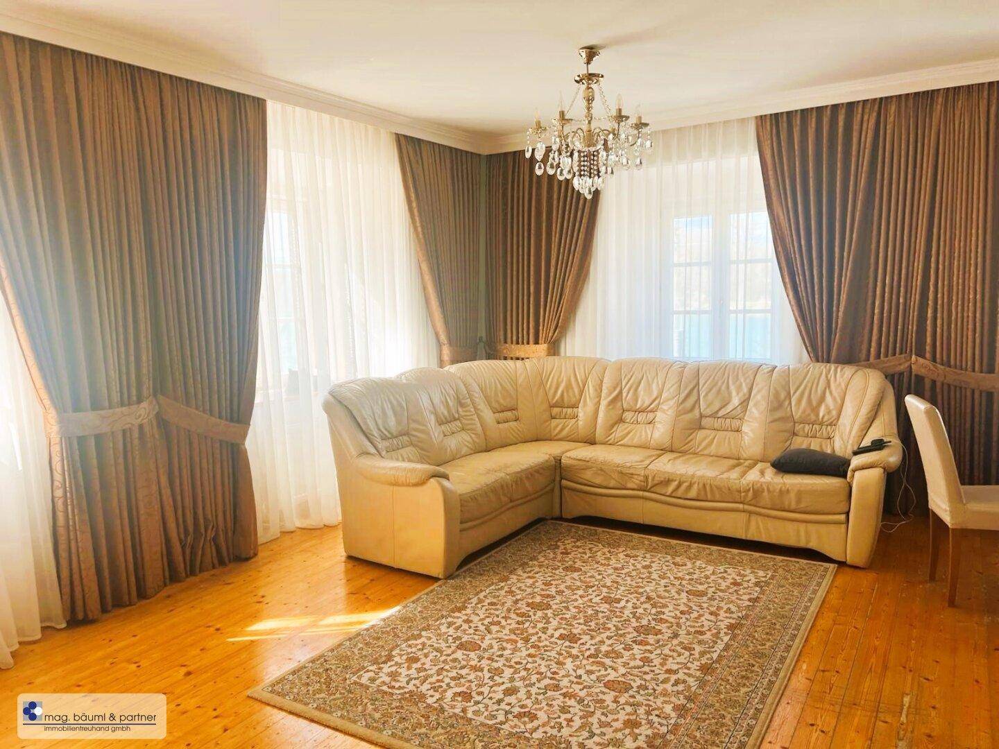 Gemütliches Wohnzimmer mit beiger Ledercouch, Parkettboden und großen Fenstern, die viel Licht hereinlassen.