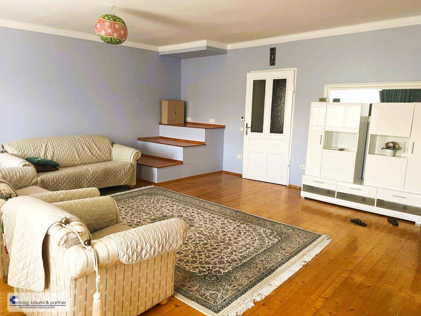 Wohnzimmer mit beigen Sofas, Parkettboden und einem Treppenaufgang, der in eine andere Etage führt.