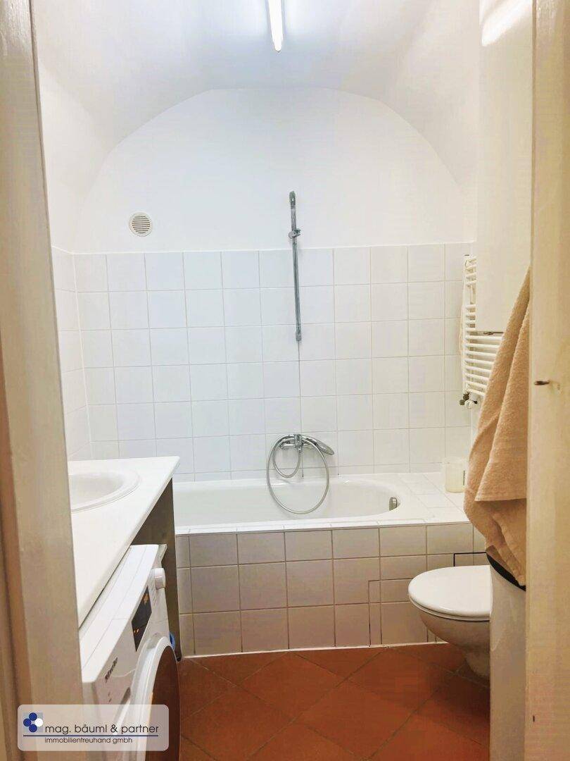 Badezimmer mit Badewanne, Toilette und beheiztem Handtuchhalter, alles in hellen Fliesen gehalten.