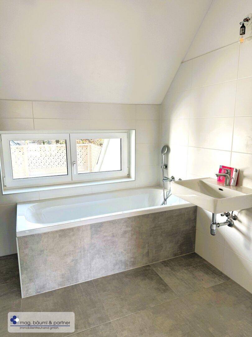 Helles Badezimmer mit Badewanne, Waschbecken und Fenster unter einer Dachschräge.