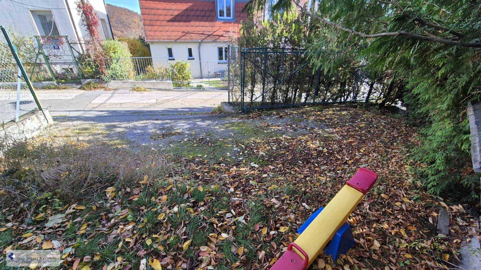 Gartenbereich mit Spielgerät und herbstlichem Laub, umgeben von Bäumen und Hecken.