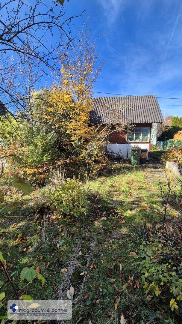 Kleines Haus mit Satteldach, umgeben von herbstlicher Vegetation und Bäumen unter blauem Himmel.