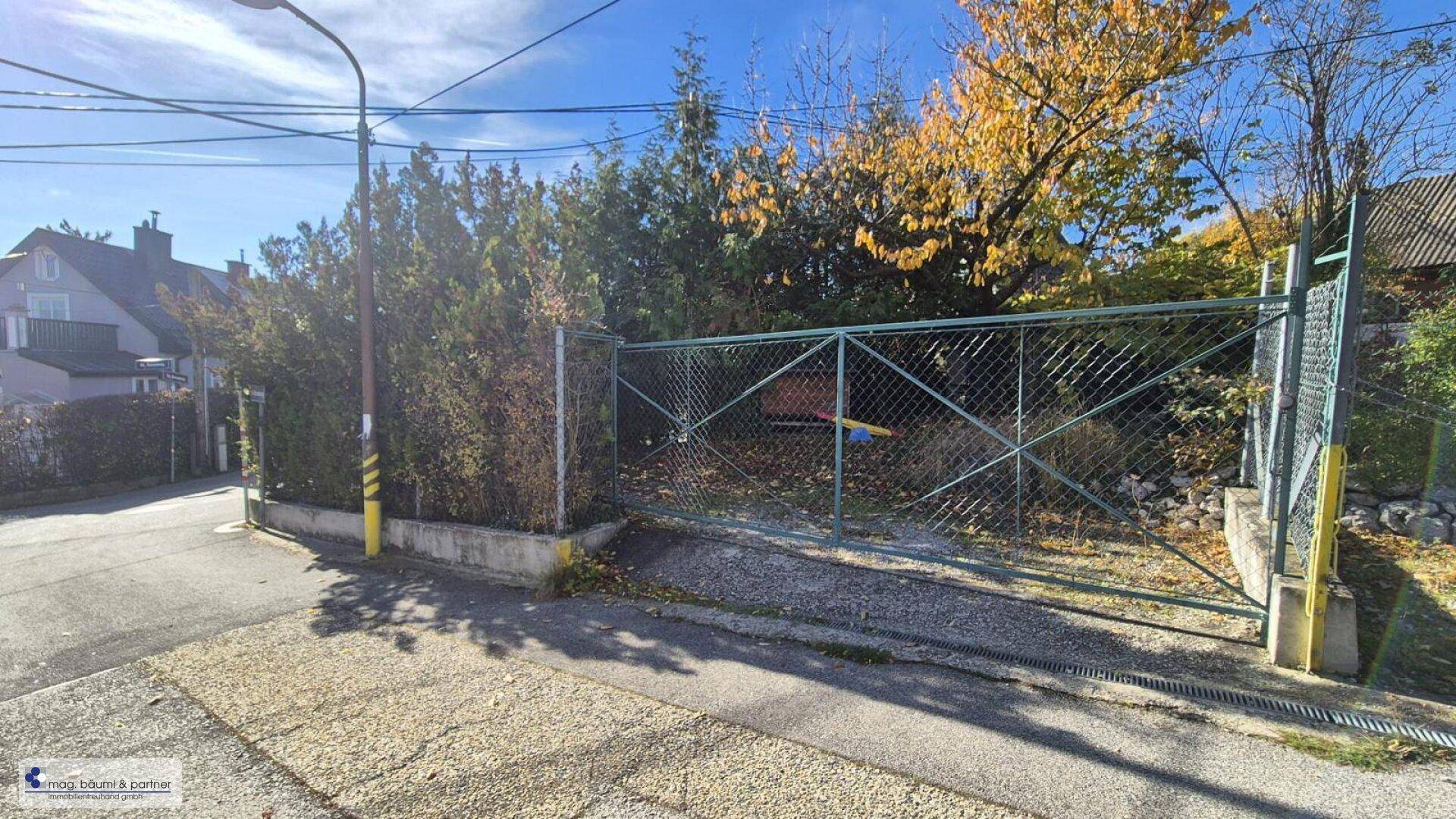Einfahrt zum Grundstück mit Metallzaun und Hecke an einer ruhigen Wohnstraße.