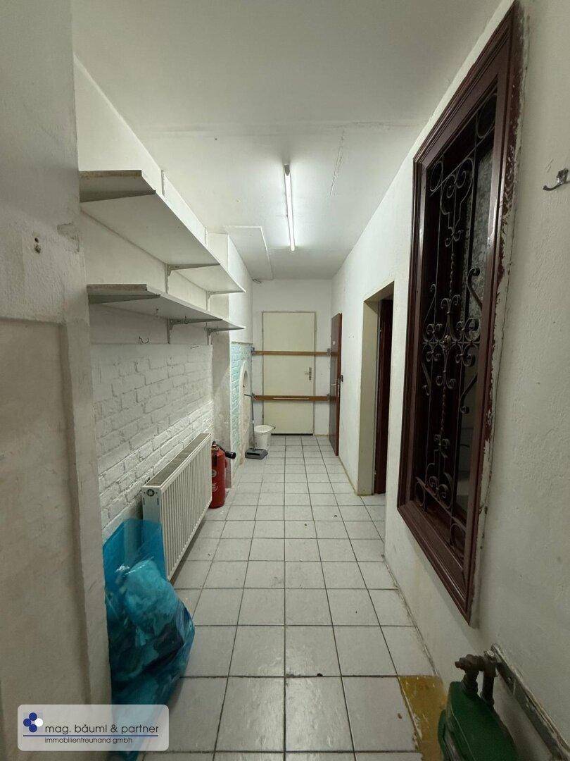 Langer, gefliester Flur mit Regalen und einem Heizkörper, der zu weiteren Räumen im Gebäude führt.