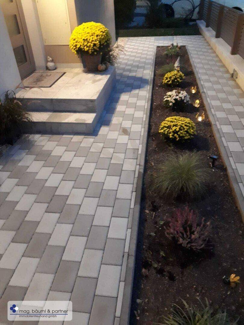 Hauseingang mit gepflastertem Weg, Blumenbeeten und dekorativen Lichtern.