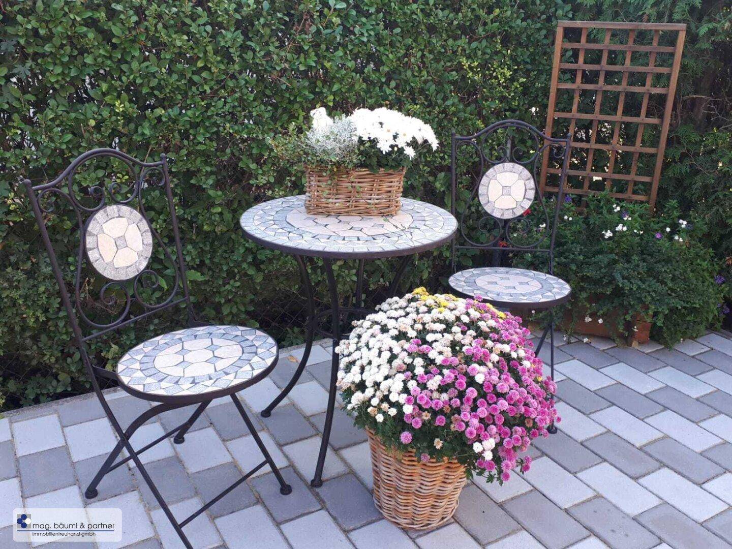 Gemütliche Sitzecke im Garten mit Mosaiktisch und Stühlen, umgeben von Blumen.