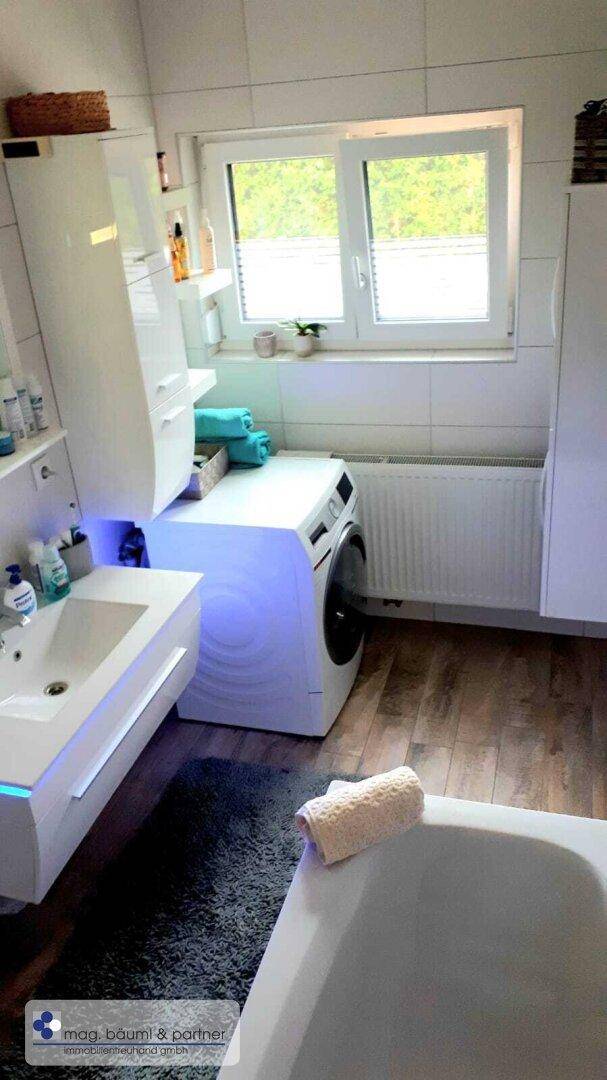 Helles Badezimmer mit Fenster, Badewanne, Waschtisch und Platz für eine Waschmaschine.