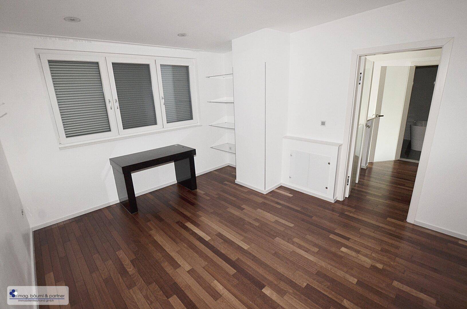 Helles Zimmer mit dunklem Holzboden, Fenster und einem modernen schwarzen Schreibtisch.