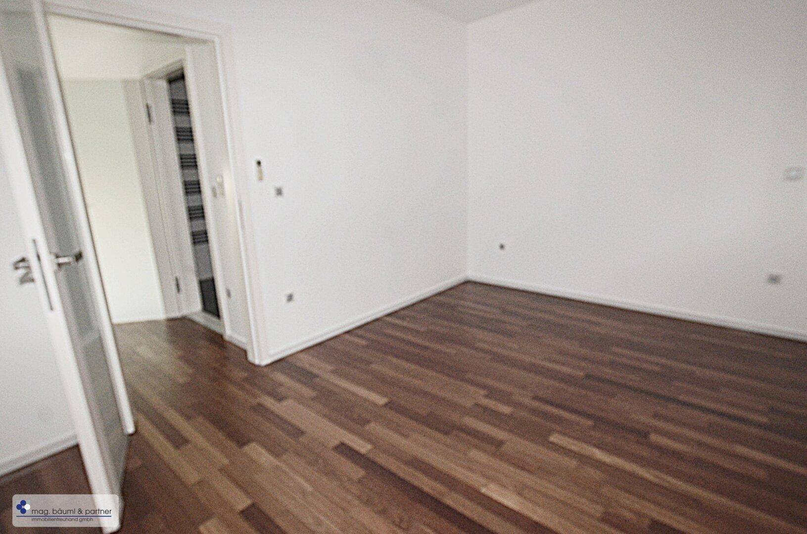 Leeres Zimmer mit dunklem Holzboden und Zugang zu einem weiteren Raum.
