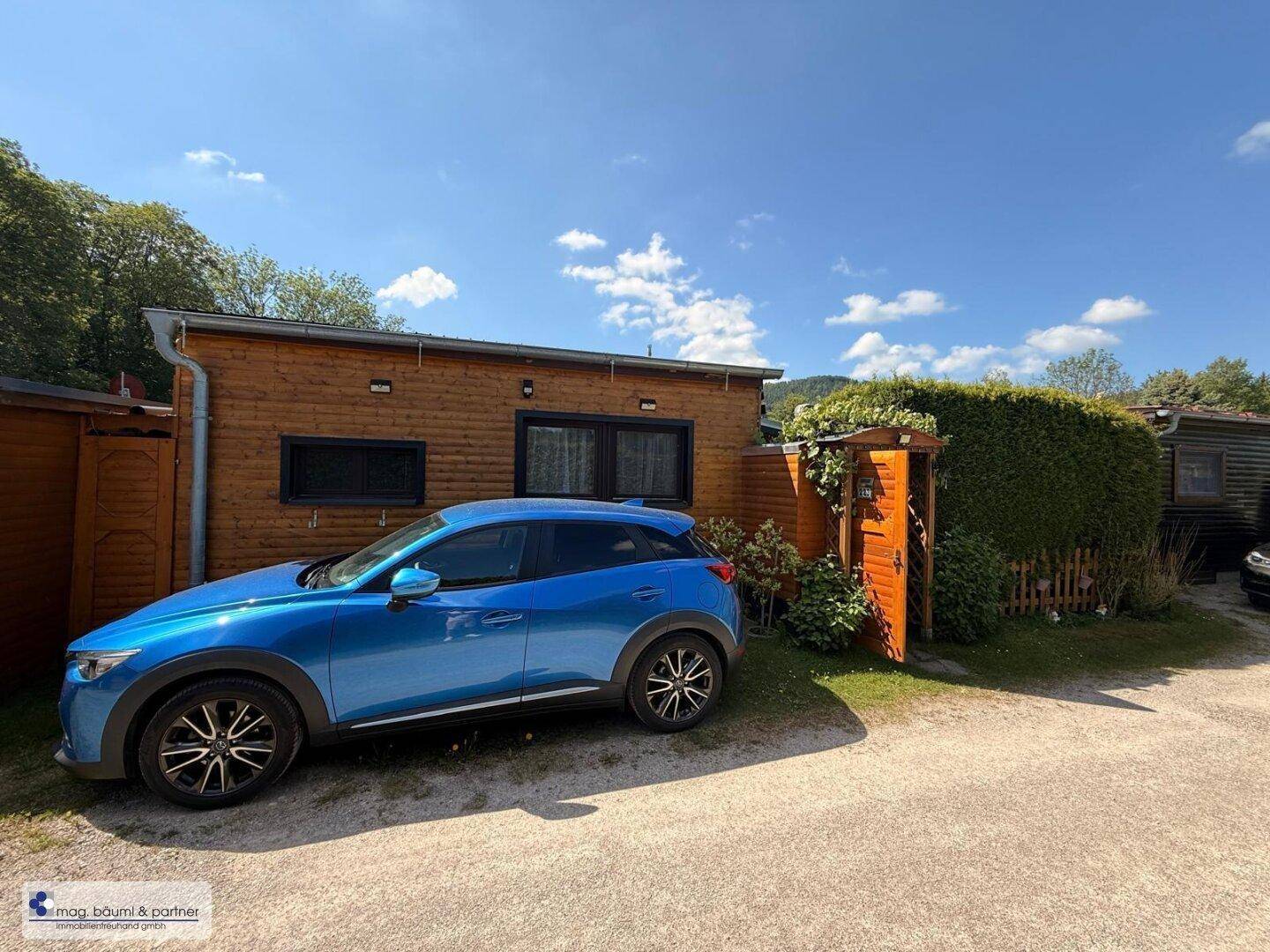 Außenansicht eines Holzhauses mit geparktem Auto und gepflegter Hecke im Vordergrund.