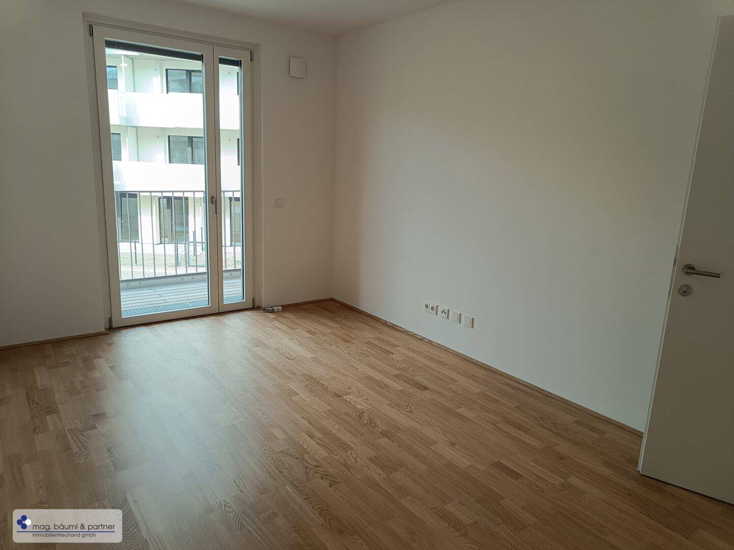 Helles Schlafzimmer mit Holzboden und Zugang zu einem Balkon durch eine große Glastür.
