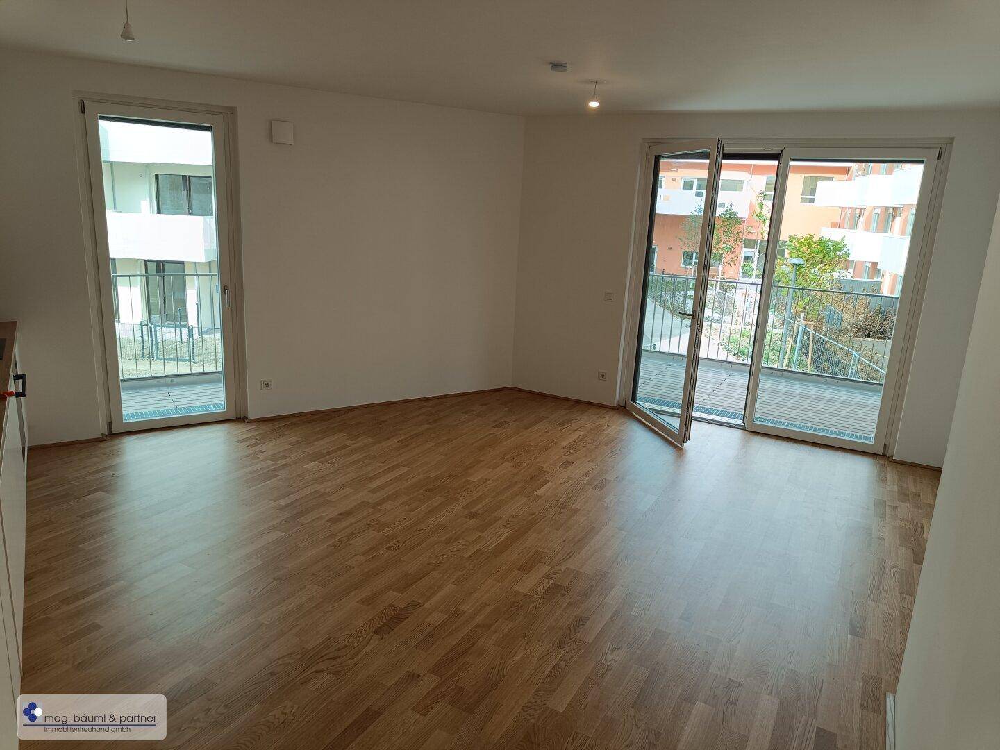 Geräumiges Wohnzimmer mit Holzboden und zwei großen Balkontüren für viel Tageslicht.