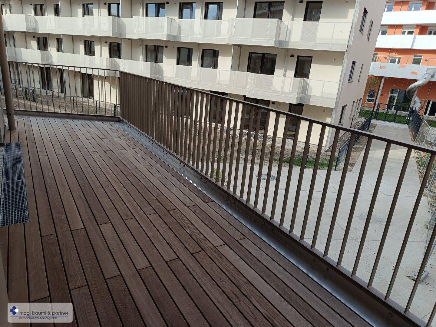 Geräumiger Balkon mit Holzbelag und Metallgeländer, bietet Blick auf die Wohnanlage.