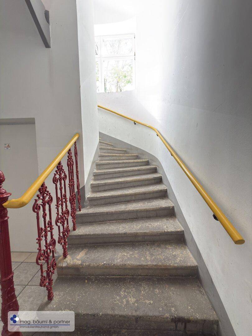 Historisches Treppenhaus mit Steinstufen, schmiedeeisernem Geländer und gelbem Handlauf.