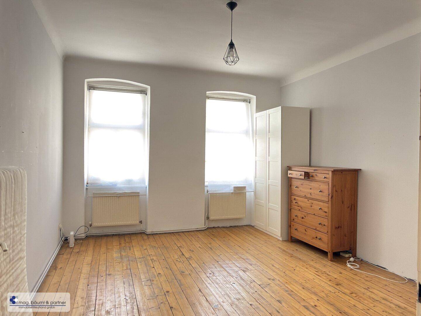 Helles Schlafzimmer mit Holzdielenboden, zwei Fenstern und weißem Kleiderschrank.