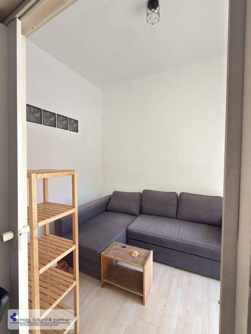 Kleines Wohnzimmer mit grauem Ecksofa und einfachem Holzregal.
