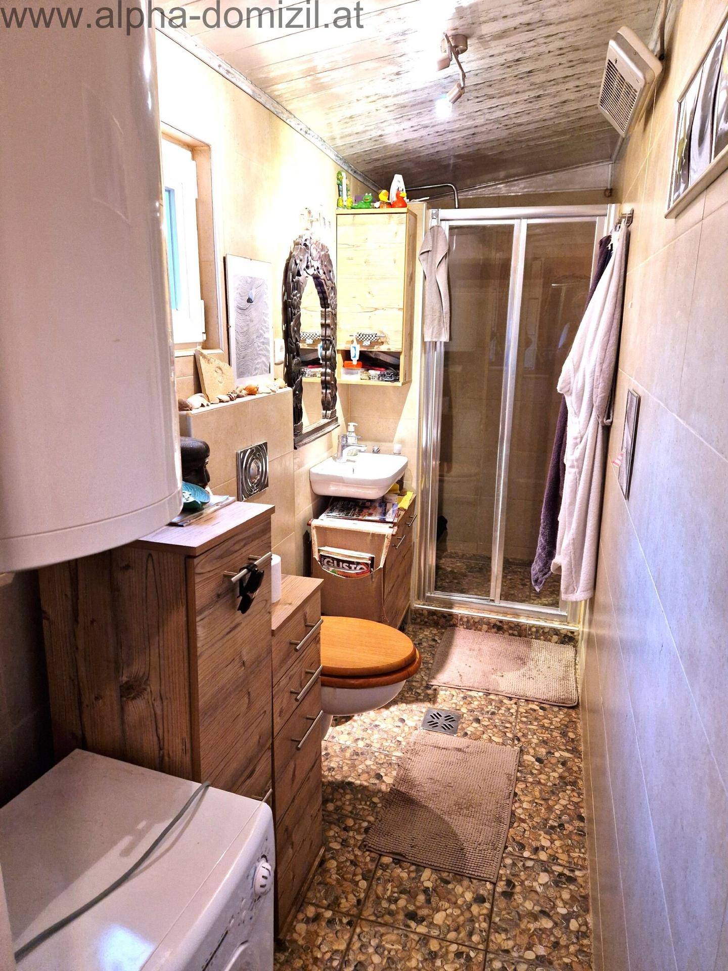 Schmales Badezimmer mit Duschkabine, WC, Waschbecken und Waschmaschine, hell gefliest.