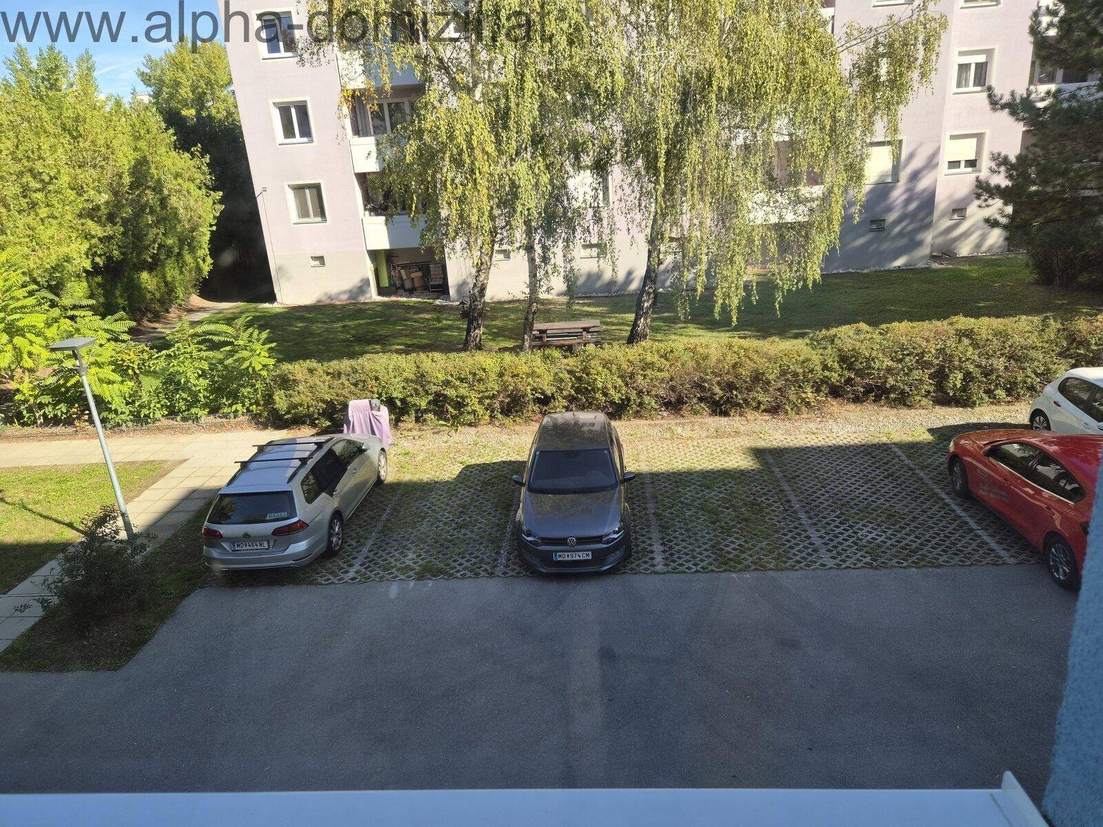 Außenansicht mit Parkplätzen, Bäumen und einem Mehrfamilienhaus im Hintergrund an einem sonnigen Tag.
