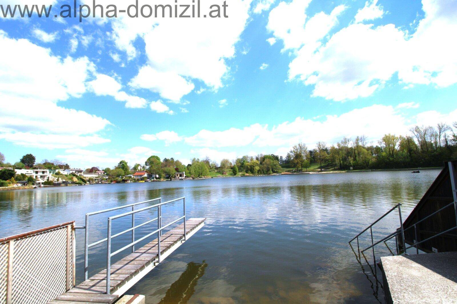 Seeufer mit Steg und Treppe ins Wasser, umgeben von grüner Landschaft und Häusern am Horizont.