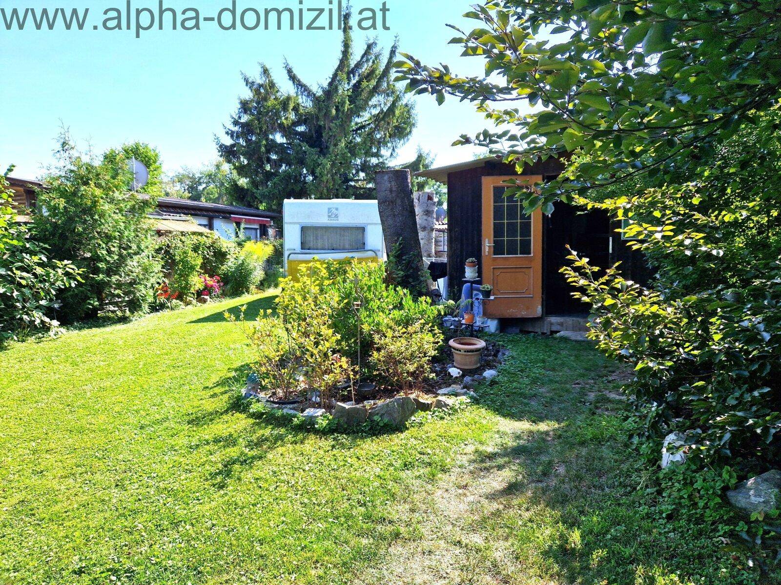 Gartenbereich mit Rasenfläche, einem kleinen Holzschuppen und einem Wohnwagen im Hintergrund.