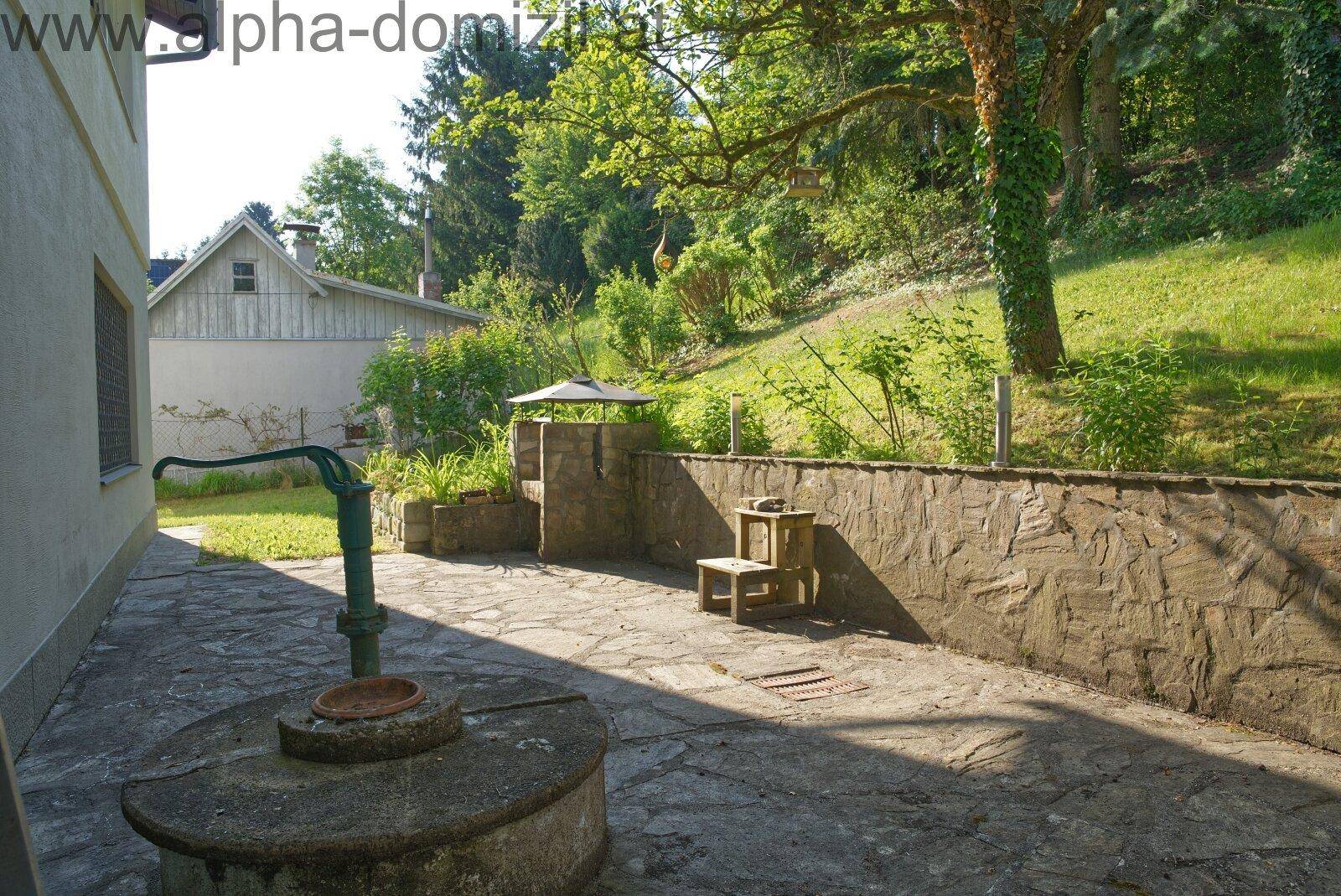 Rustikale Terrasse mit Steinmauer und einem alten Brunnen, umgeben von viel Grün.