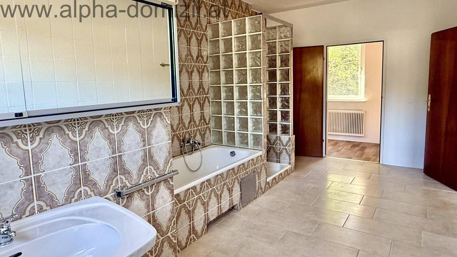 Badezimmer im 80er-Jahre-Stil mit gemusterten Fliesen, Badewanne und Glasbausteinwand, sowie einem Waschbecken.