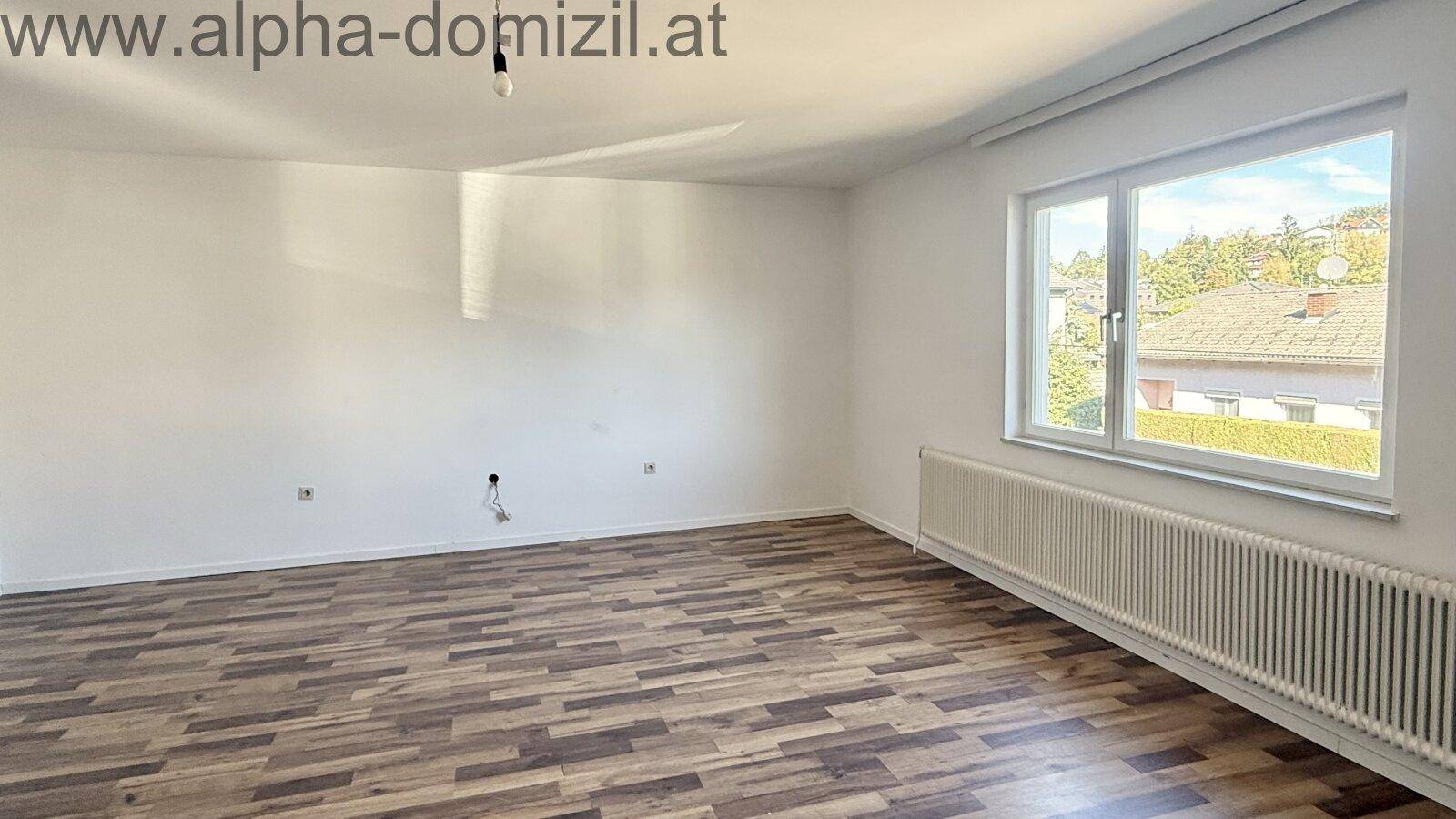 Heller, leerer Raum mit Laminatboden und einem großen Fenster, das viel Tageslicht hereinlässt.