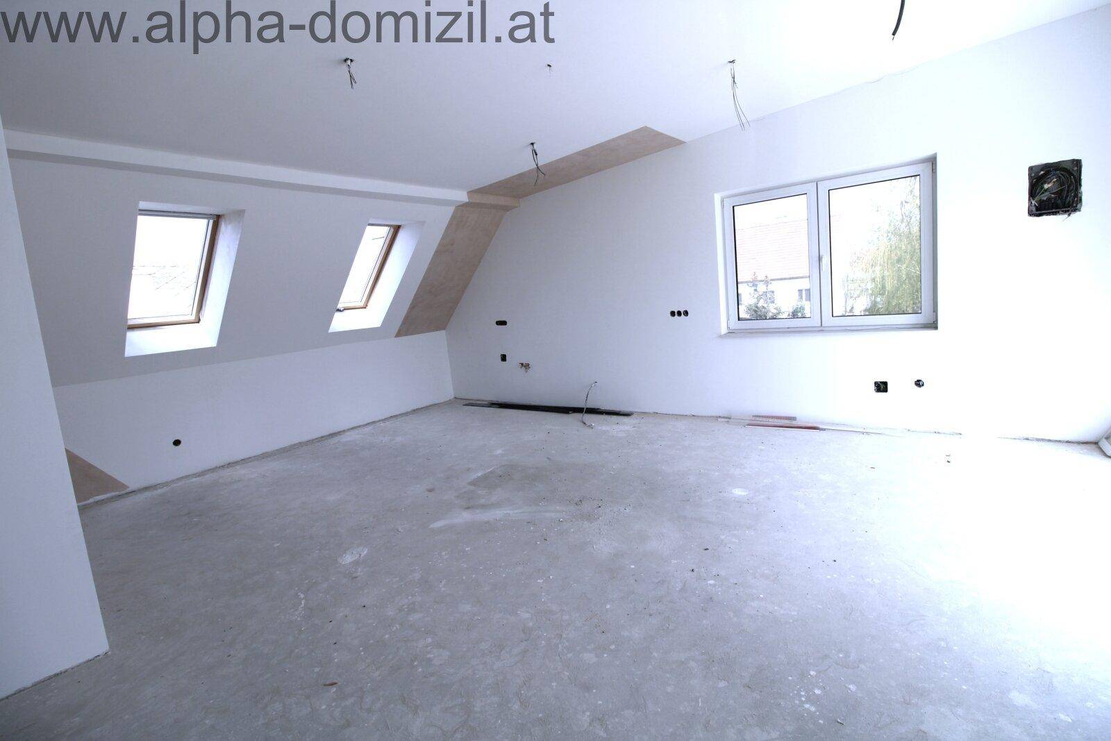 Geräumiges Dachzimmer mit schrägen Wänden, Dachfenstern und Rohbetonboden.