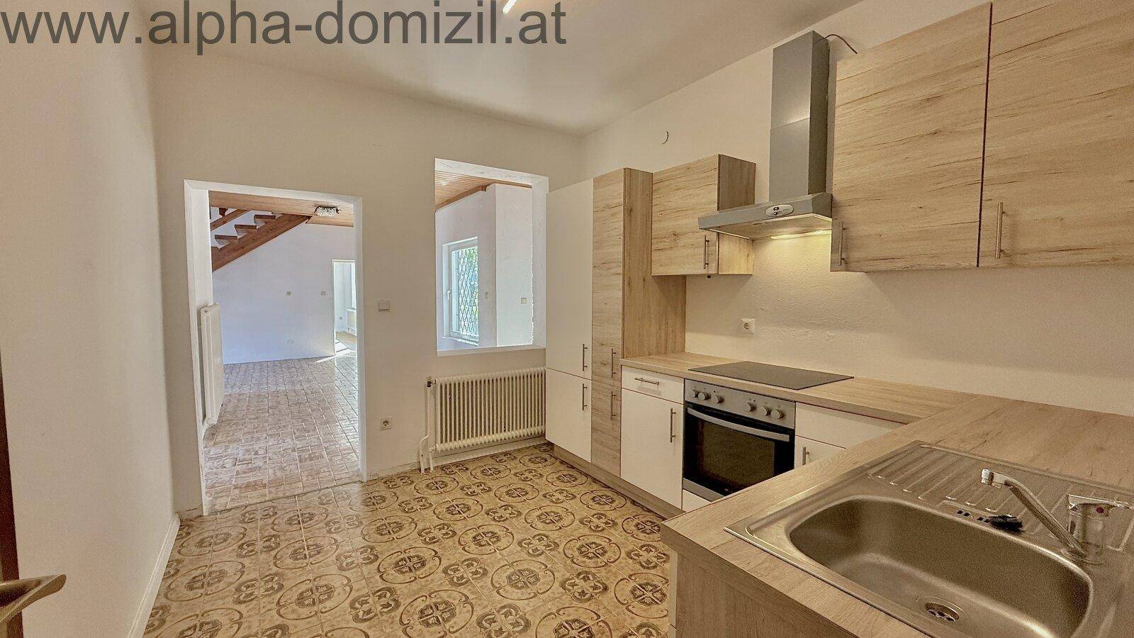 Funktionale Küche mit Holzschränken, Spüle, Herd und Fliesenboden, mit Blick in einen weiteren Raum.