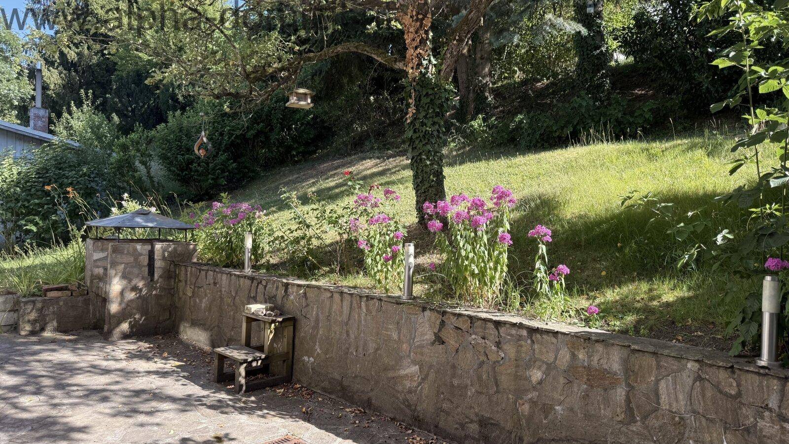 Gepflegter Gartenbereich mit Steinmauer, blühenden Pflanzen und Rasenfläche unter Bäumen.
