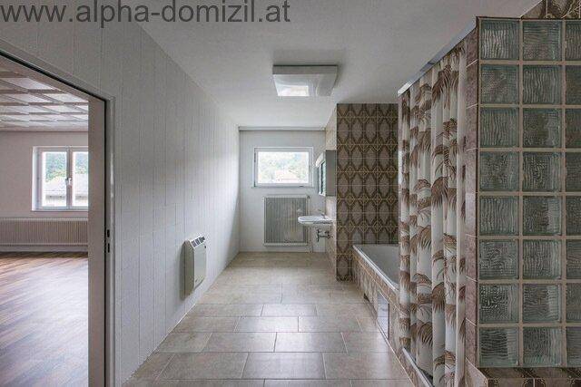 Langes Badezimmer mit Badewanne, Waschbecken und Fenster, gefliest im 80er-Jahre-Stil.
