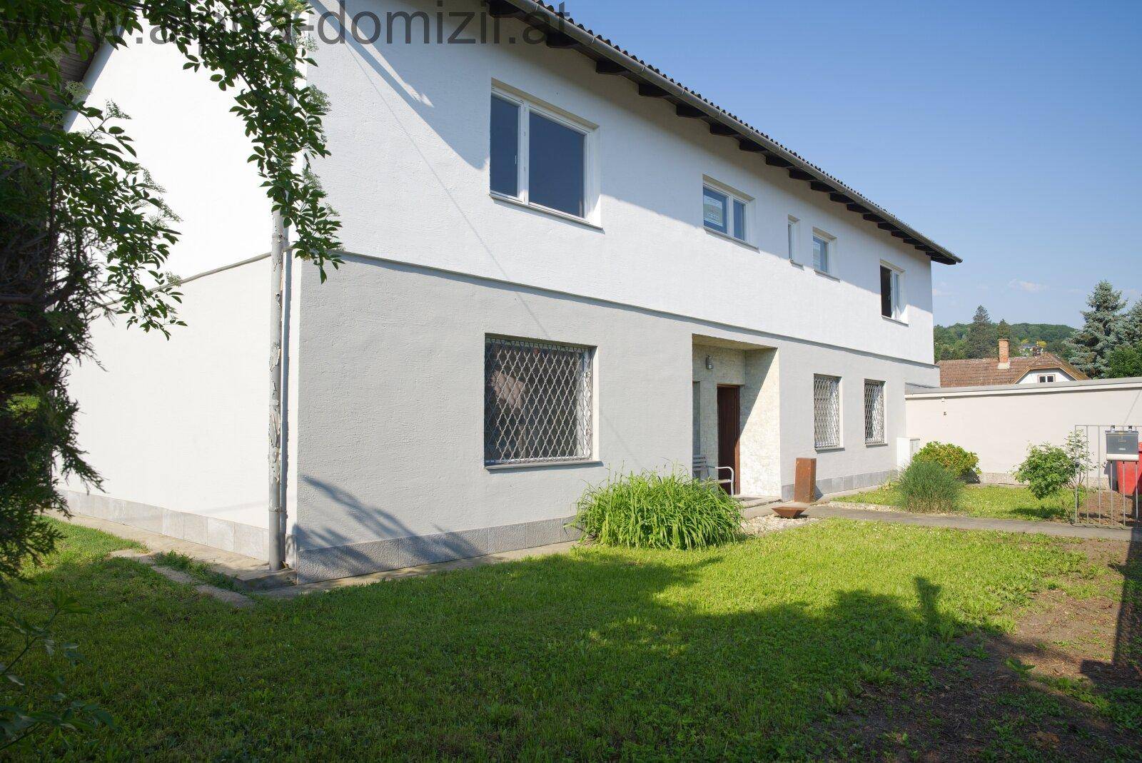 Seitenansicht des Hauses mit weißer Fassade, Rasenfläche und Fenstern mit Gitterrosten.