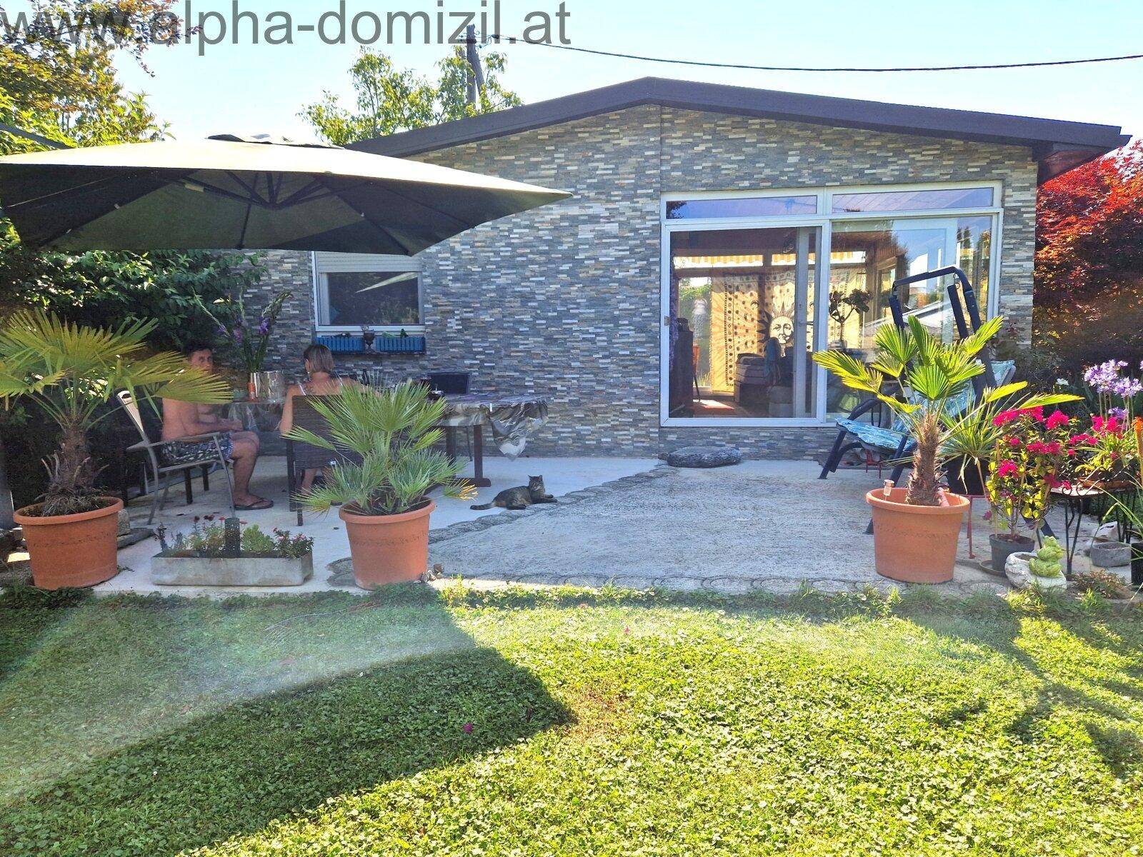 Gemütliche Terrasse mit Sitzgelegenheiten, Sonnenschirm und Topfpflanzen vor einem kleinen Haus.