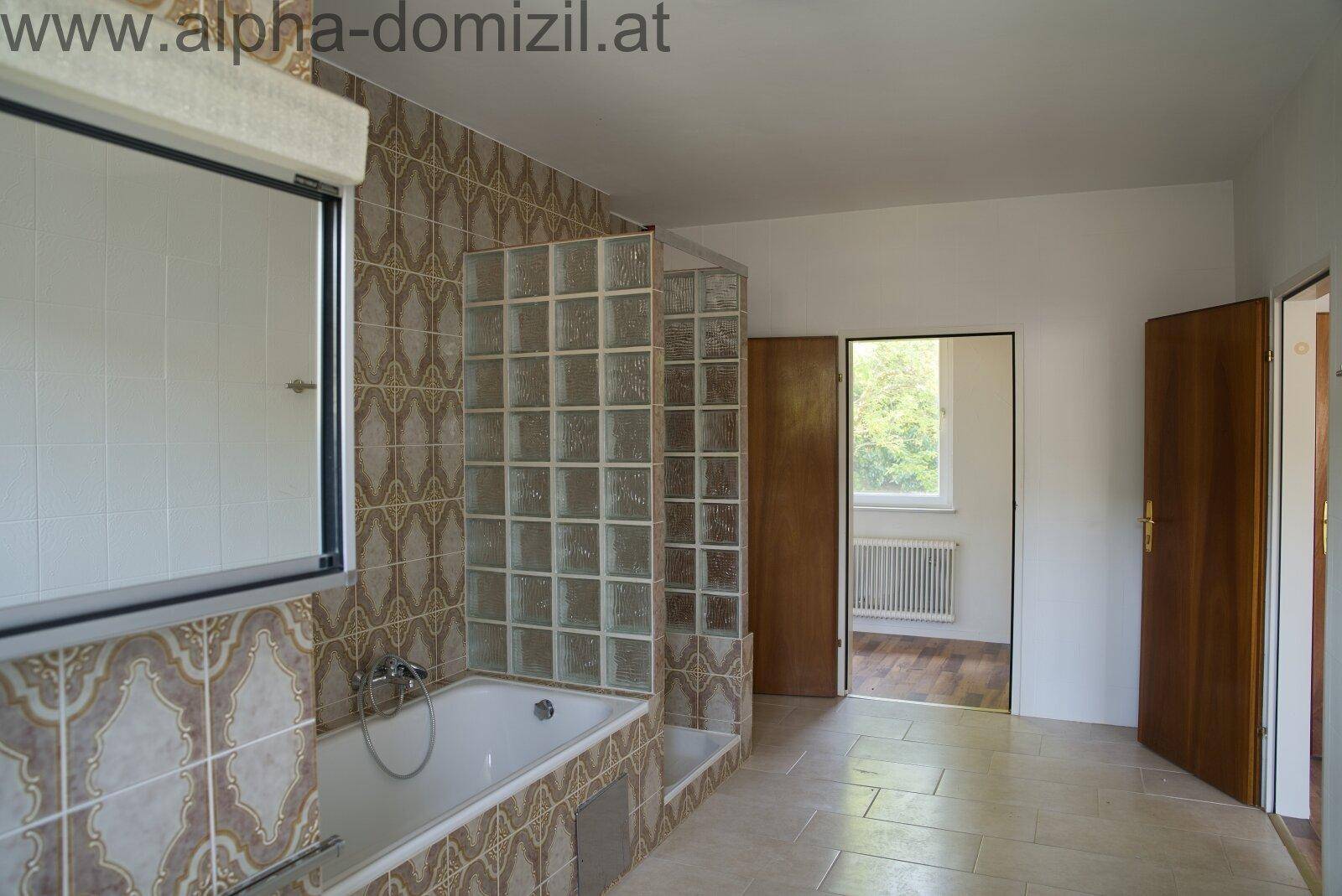 Badezimmer mit Badewanne, Glasbaustein-Duschabtrennung und gemusterten Fliesen im 80er-Jahre-Stil.