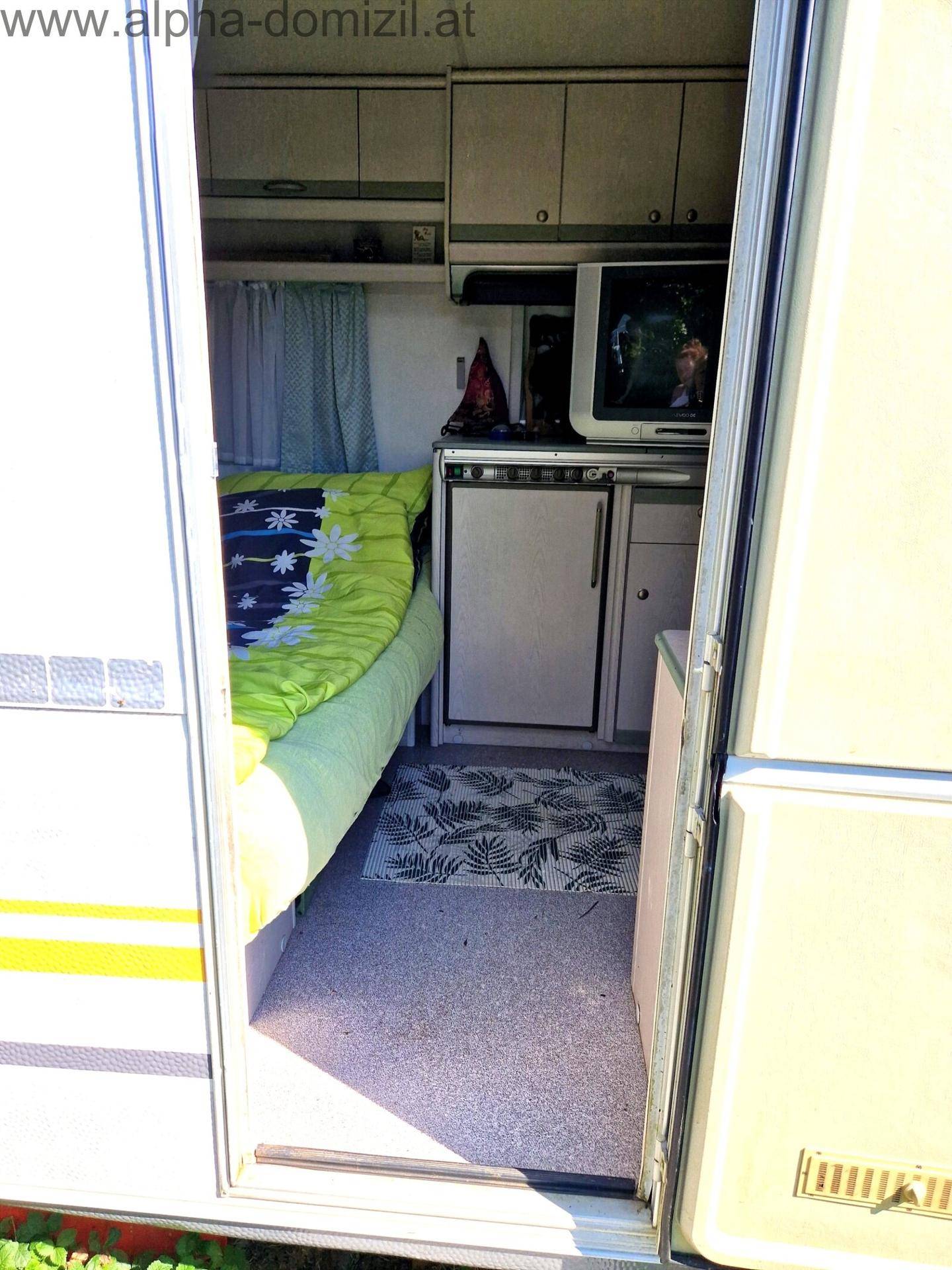 Innenansicht eines Wohnwagens mit Schlafbereich, kleinem Kühlschrank und Stauraum.