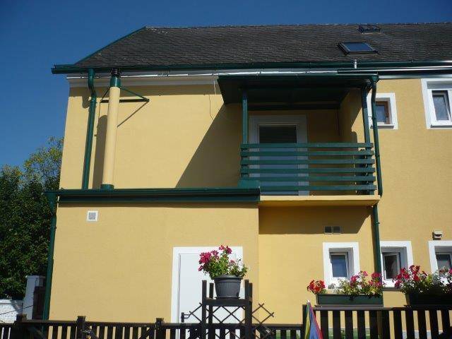 Kleiner Balkon an der gelben Fassade mit grünen Geländern und blühenden Blumenkästen in der Sonne.