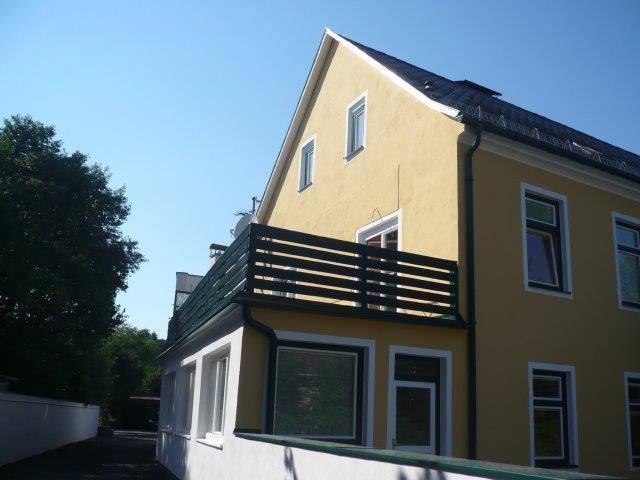 Seitliche Ansicht des Gebäudes mit großem Balkon und Blick ins Grüne unter blauem Himmel.