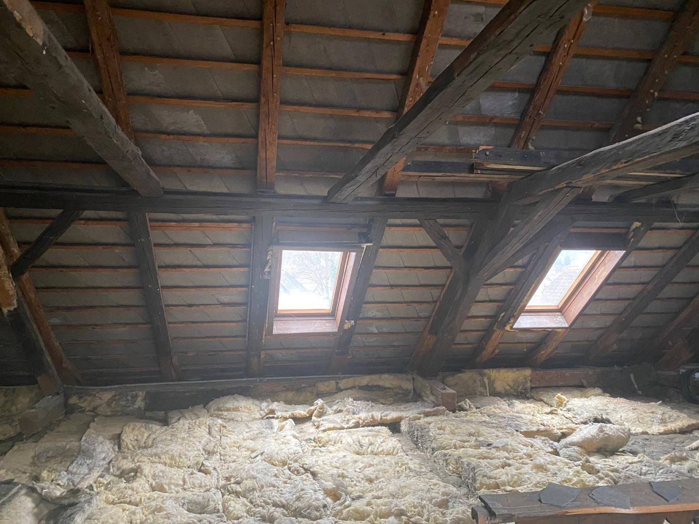 Dachboden im Rohzustand mit sichtbaren Holzbalken, Isolierung und zwei Dachfenstern für natürliches Licht.