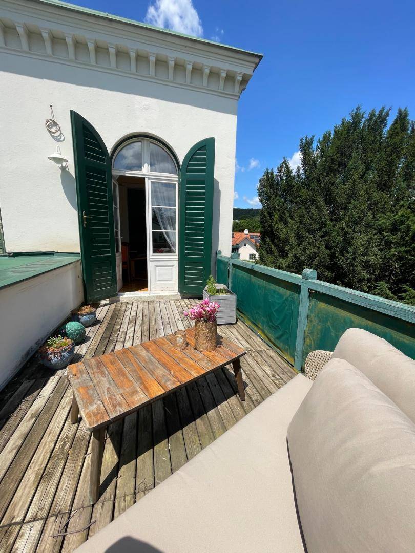 Sonniger Balkon mit Holzboden, Sitzgelegenheiten und Blick ins Grüne unter blauem Himmel.