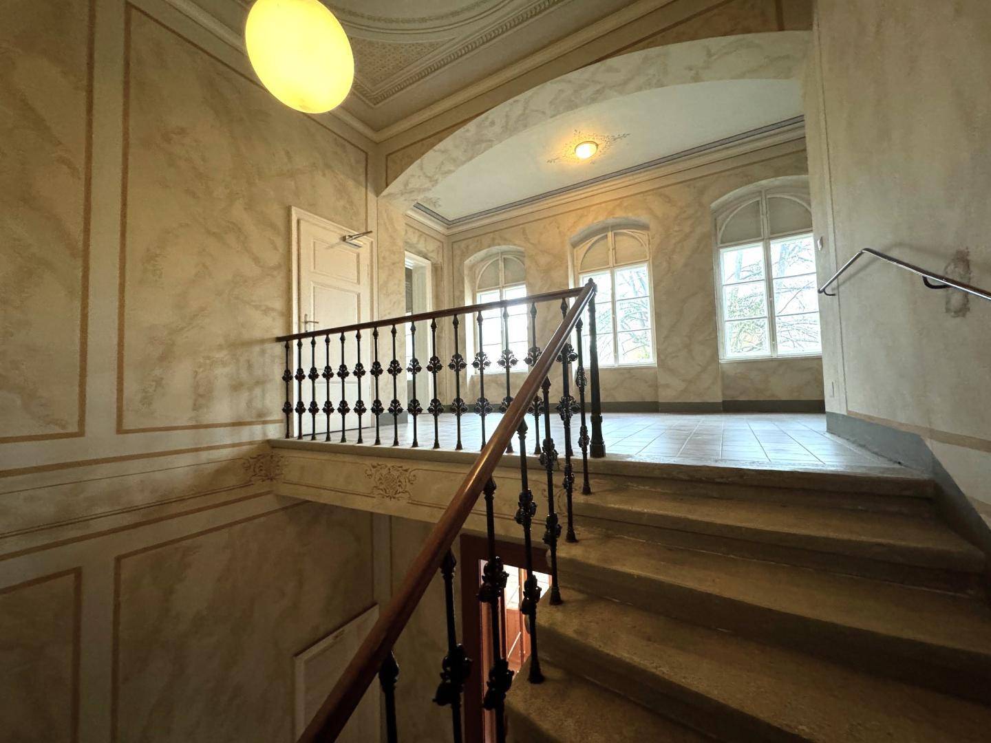 Elegantes Treppenhaus mit kunstvollem Geländer, Marmoroptik an den Wänden und hohen Bogenfenstern.