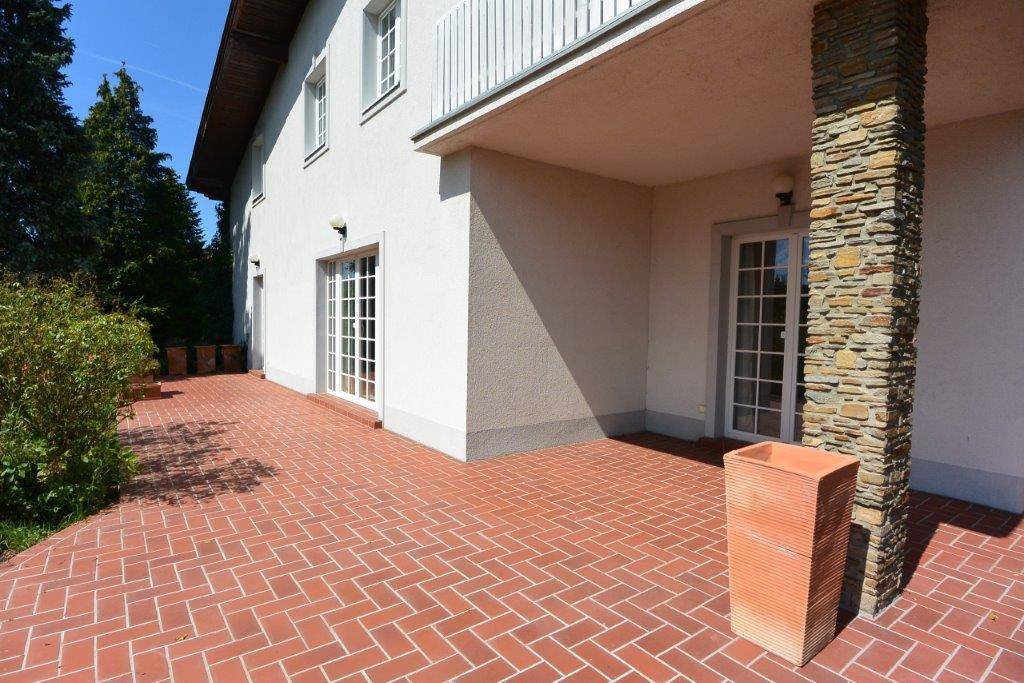 Geräumige Ziegelterrasse mit Steinsäule und Zugangstüren zum Haus, umgeben von Grün.