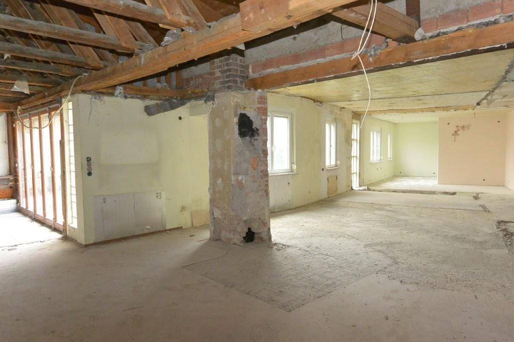Großer Innenraum im Rohbau mit freiliegenden Holzbalken und mehreren Fenstern, bereit zur Neugestaltung.