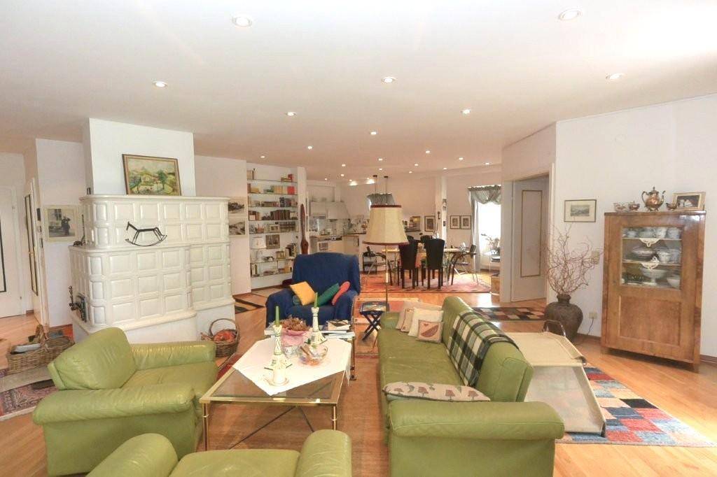 Heller Wohnbereich mit Kachelofen und grünen Sofas, offen gestaltet zum Essbereich und zur Küche im Hintergrund.