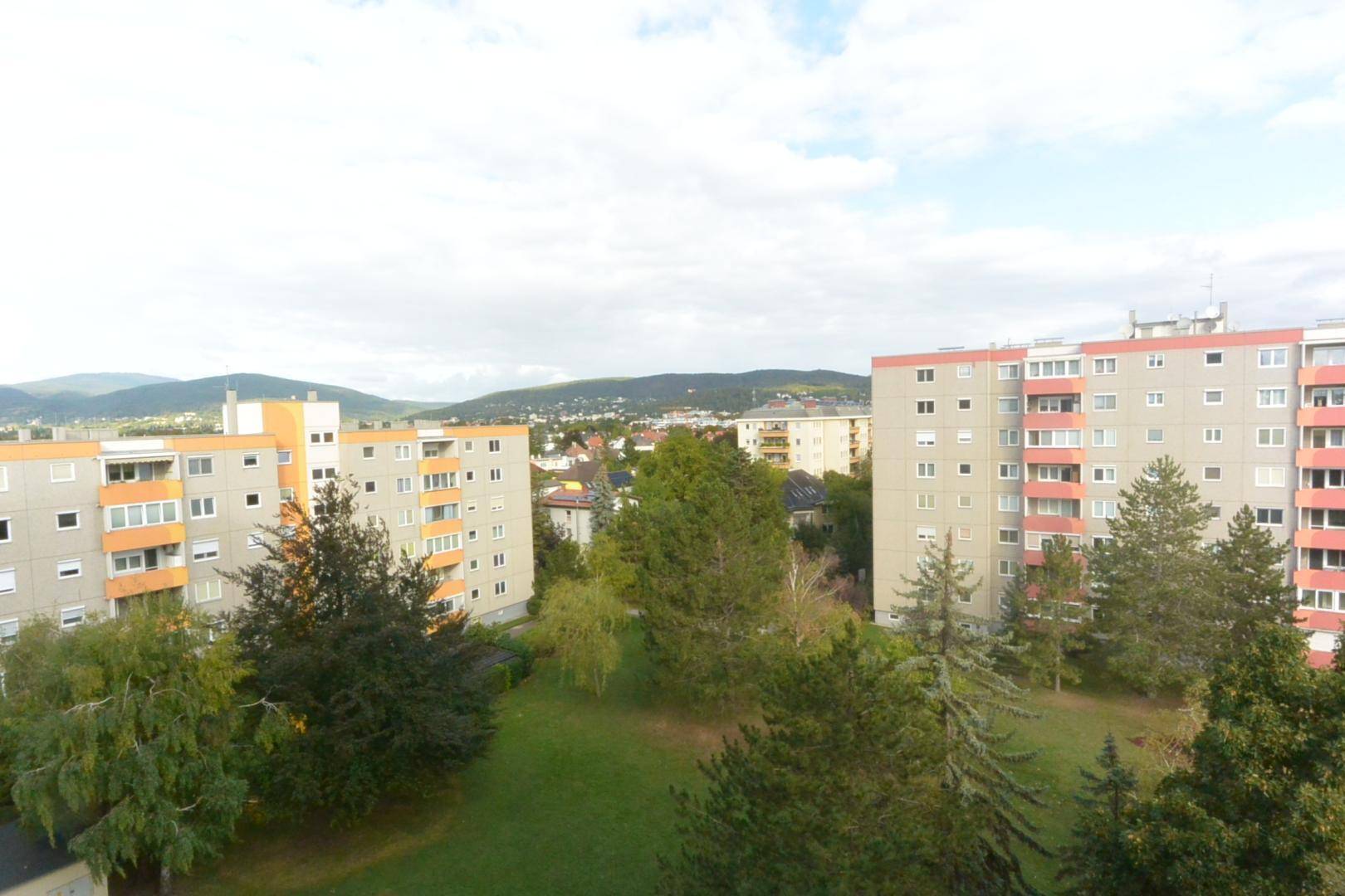 Blick auf Wohngebäude mit Grünflächen und bewaldeten Hügeln im Hintergrund.
