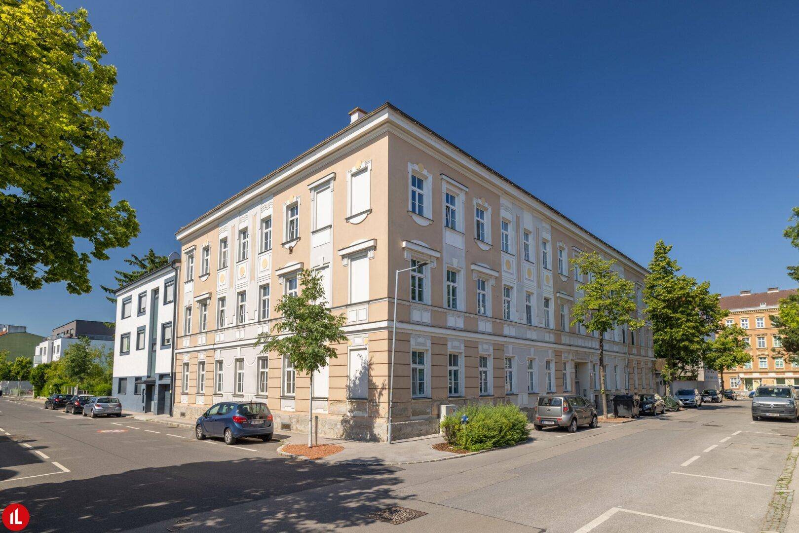 Eckansicht eines gepflegten Altbaus mit beiger Fassade und vielen Fenstern an einer sonnigen Straße.