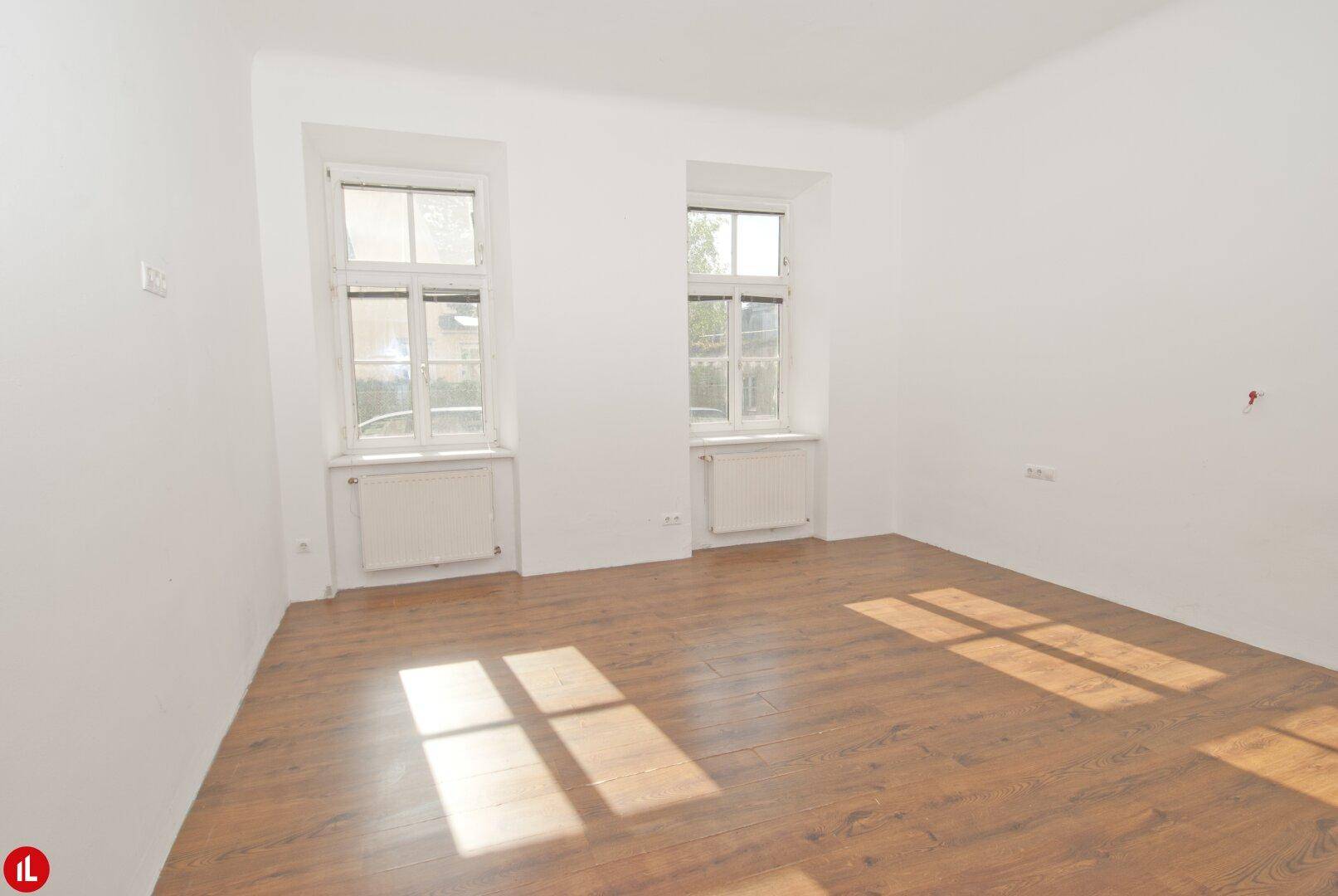 Leeres Zimmer mit hellem Laminatboden und zwei großen Fenstern, durch die Sonnenlicht fällt.
