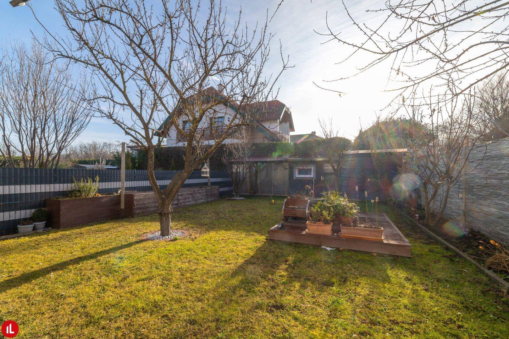 Sonniger Gartenbereich mit Rasenfläche, Bäumen und einer kleinen Holzterrasse.