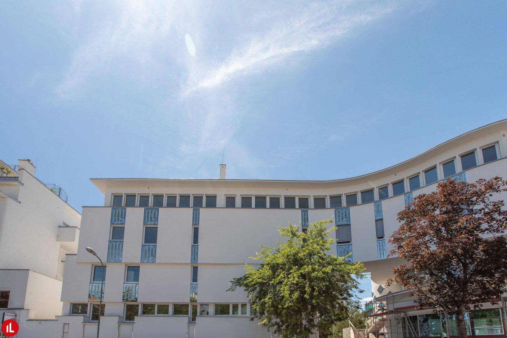 Ansicht eines modernen weißen Gebäudes mit geschwungener Fassade und vielen Fenstern, umgeben von Bäumen.